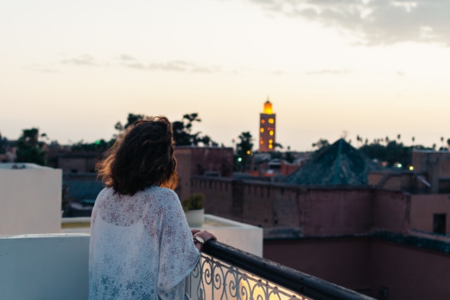 Žena v bielych čipkovaných šatách stojí pri zábradlí na balkóne počas východu slnka